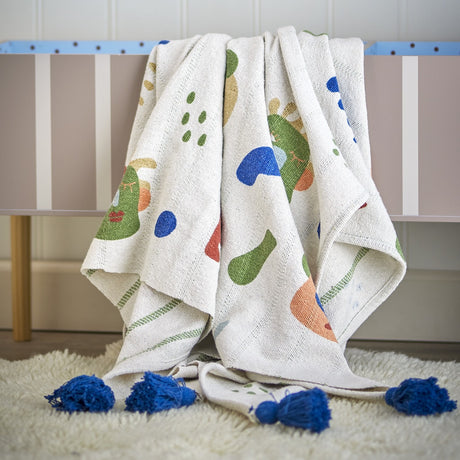 Sylvie Cotton Throw in multicoloured abstract print with blue tassels, draped over a cot rail with a cream sheepskin rug beneath.