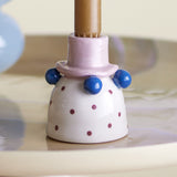 Tacana Dotted Stoneware Candle Holder in white with burgundy spots, featuring a pink hat-shaped socket and blue glossy beads, holding a brown candle.