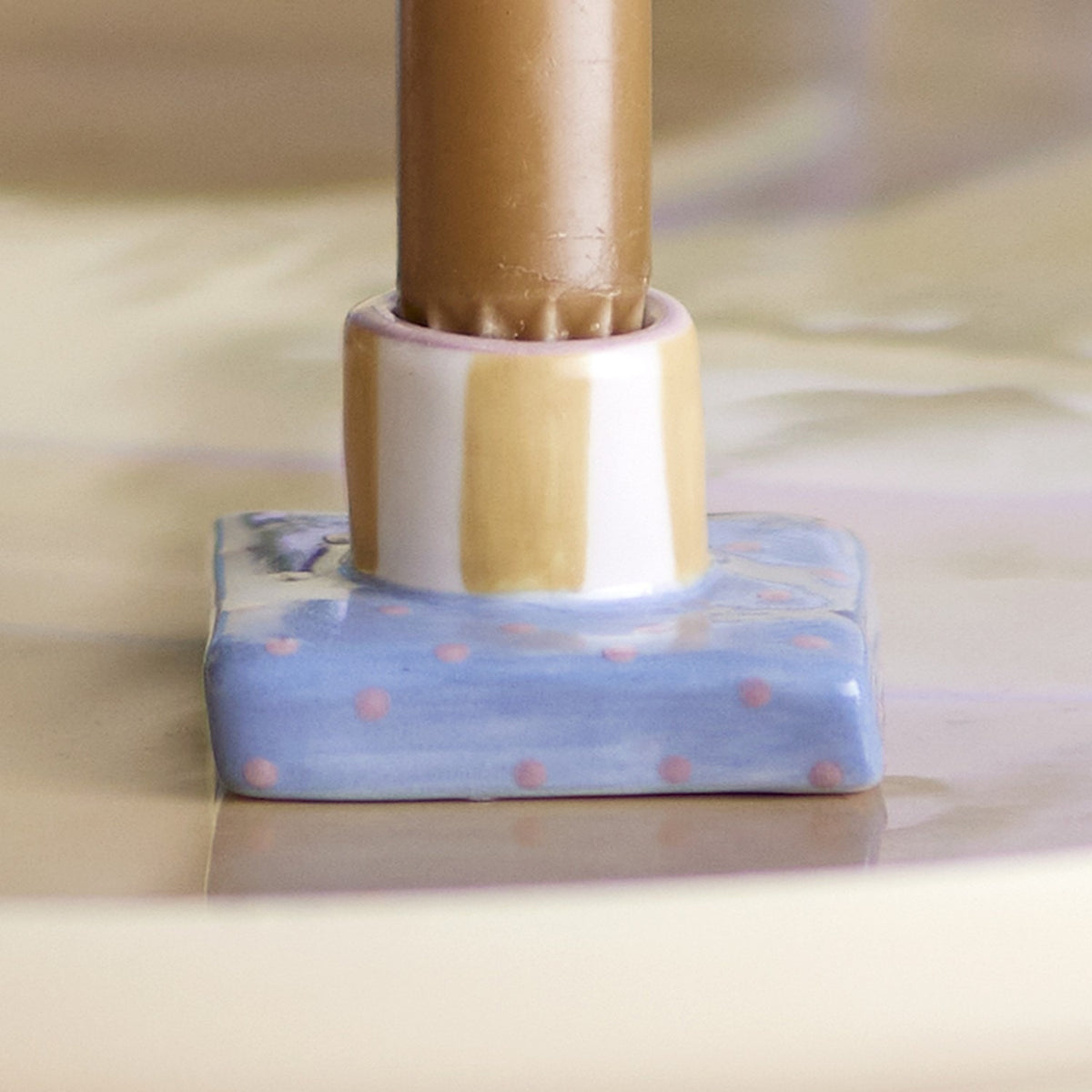 Close-up of the Tacana Striped Stoneware Candle Holder, showing a white and tan striped round cup on a blue square base with pink dots, holding a wooden candle.