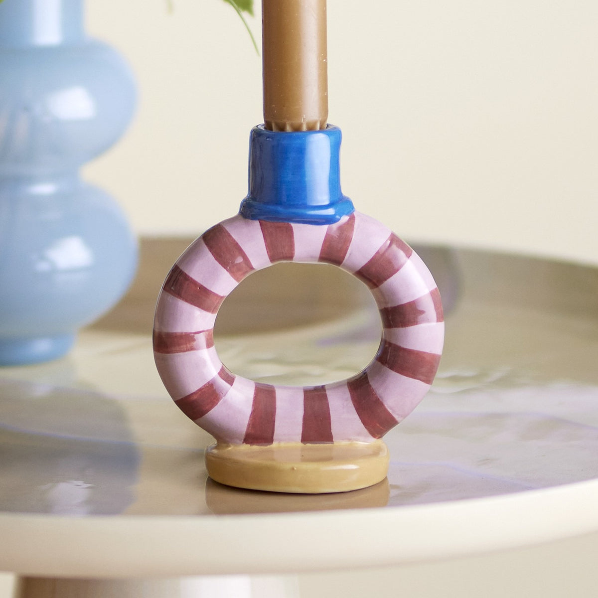 Tacana Striped Stoneware Candle Holder, 12cm, open ring shape with pink and brown stripes, blue cylindrical socket holding a taper candle, round beige base, shown on a tabletop.