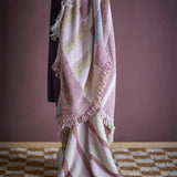 Lane Striped Throw in multicoloured woven cotton blend with wide block stripes and long fringed edges, draped over a chair against a dark pink wall.