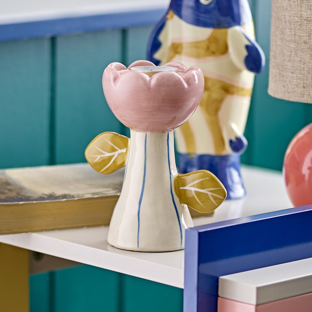 Florita Flower Tealight & Candle Holder in handpainted ceramic, pink scalloped petal cup on a striped white stem with mustard leaves either side, shown on a shelf with decor.