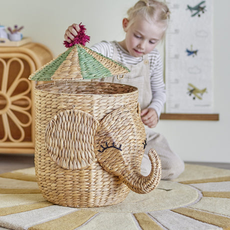 Bobo Elephant water hyacinth storage basket with trunk, ears and stitched eyelashes, topped by a green striped conical lid with pink pompom, shown with a child lifting the lid indoors.