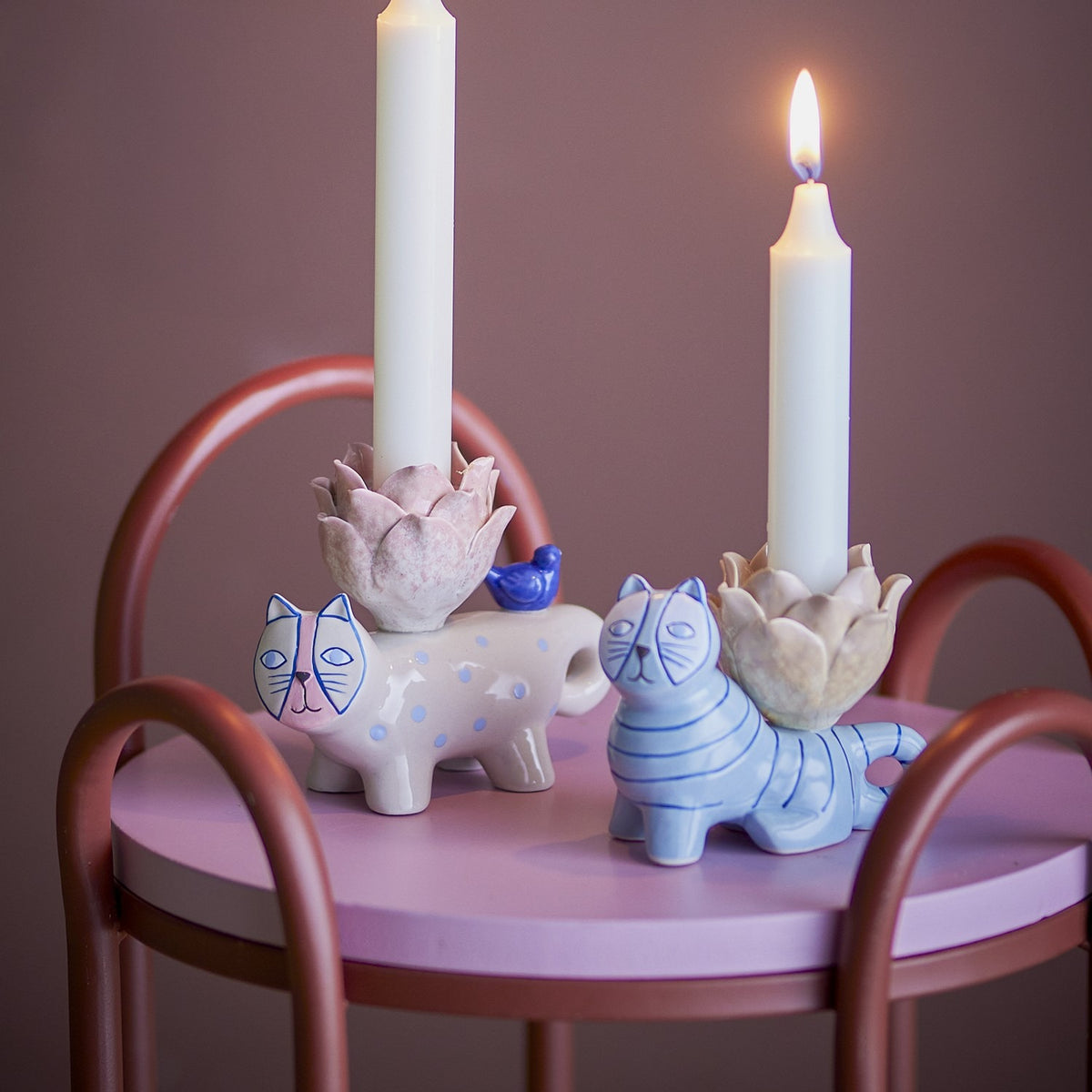 Missy Cat Stoneware Candle Holders set of two, a spotted beige cat with a blue bird and a striped blue cat, each with a flower-shaped cup holding a taper candle on a pink table.