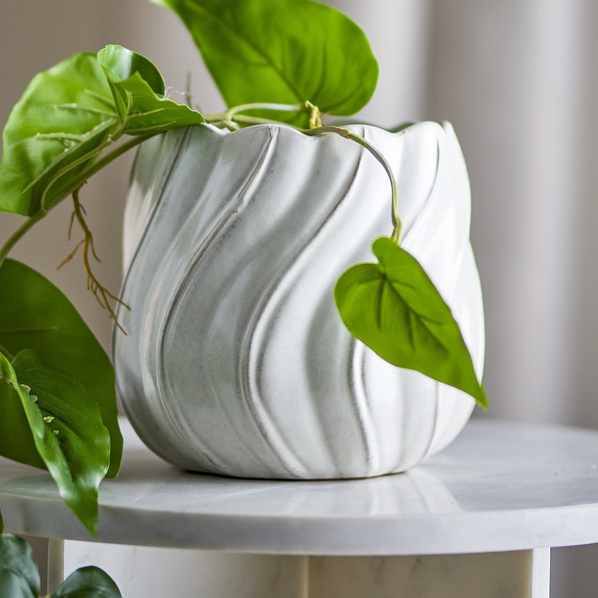 Isidora Wavy Stoneware Plant Pot in White, with scalloped rim and diagonal wave texture, shown on a marble side table with trailing green foliage.