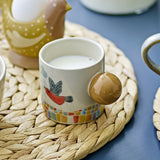 Colorine Stoneware Cup 155ml in multicoloured ceramic, with a round ball handle, painted bird and cloud design, and a mosaic patterned base, filled with milk on a woven mat.