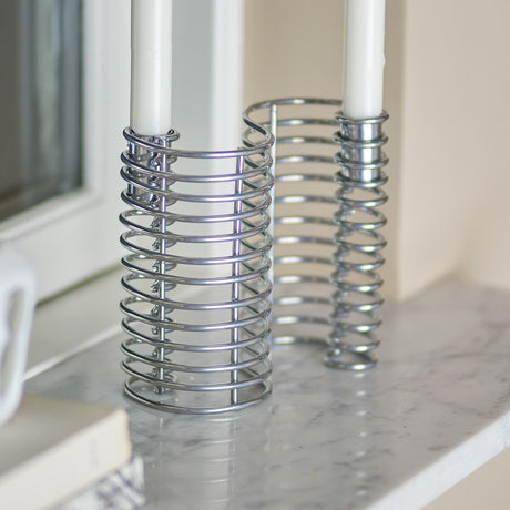 Wavela Wavy Iron Candle Holder in silver, an S-shaped coiled wire piece holding two white taper candles, shown on a marble windowsill ledge.