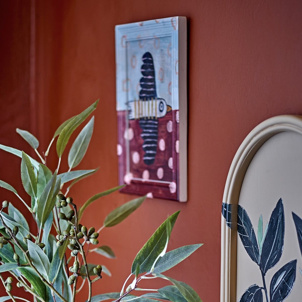 Brett Birdy Wooden Framed Print in multicoloured tones, showing a striped bird design with scalloped wings, rings and dots, mounted on a terracotta wall beside foliage.