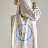 Beige cotton tote bag with confetti print, carried over the shoulder, showing a scalloped blue roundel with "Bloomingville mini" wordmark on the front.