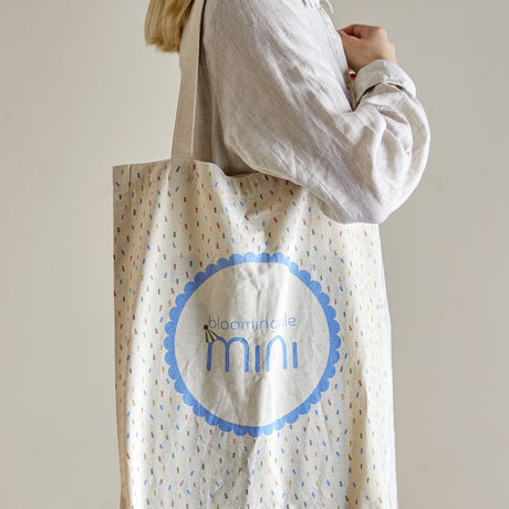 Beige cotton tote bag with confetti print, carried over the shoulder, showing a scalloped blue roundel with "Bloomingville mini" wordmark on the front.