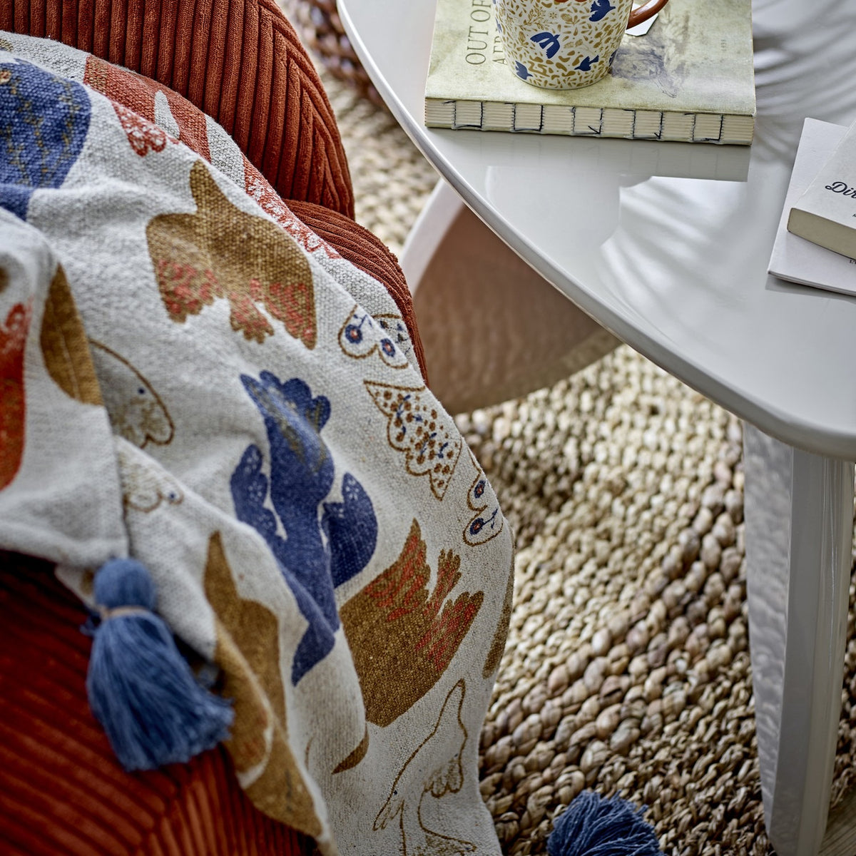 Anniversary Cotton Throw in multicoloured bird and wing print with blue tassel corners, draped over a rust corduroy armchair beside a white side table.