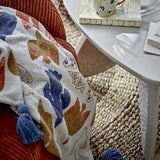 Anniversary Cotton Throw in multicoloured bird and wing print with blue tassel corners, draped over a rust corduroy armchair beside a white side table.