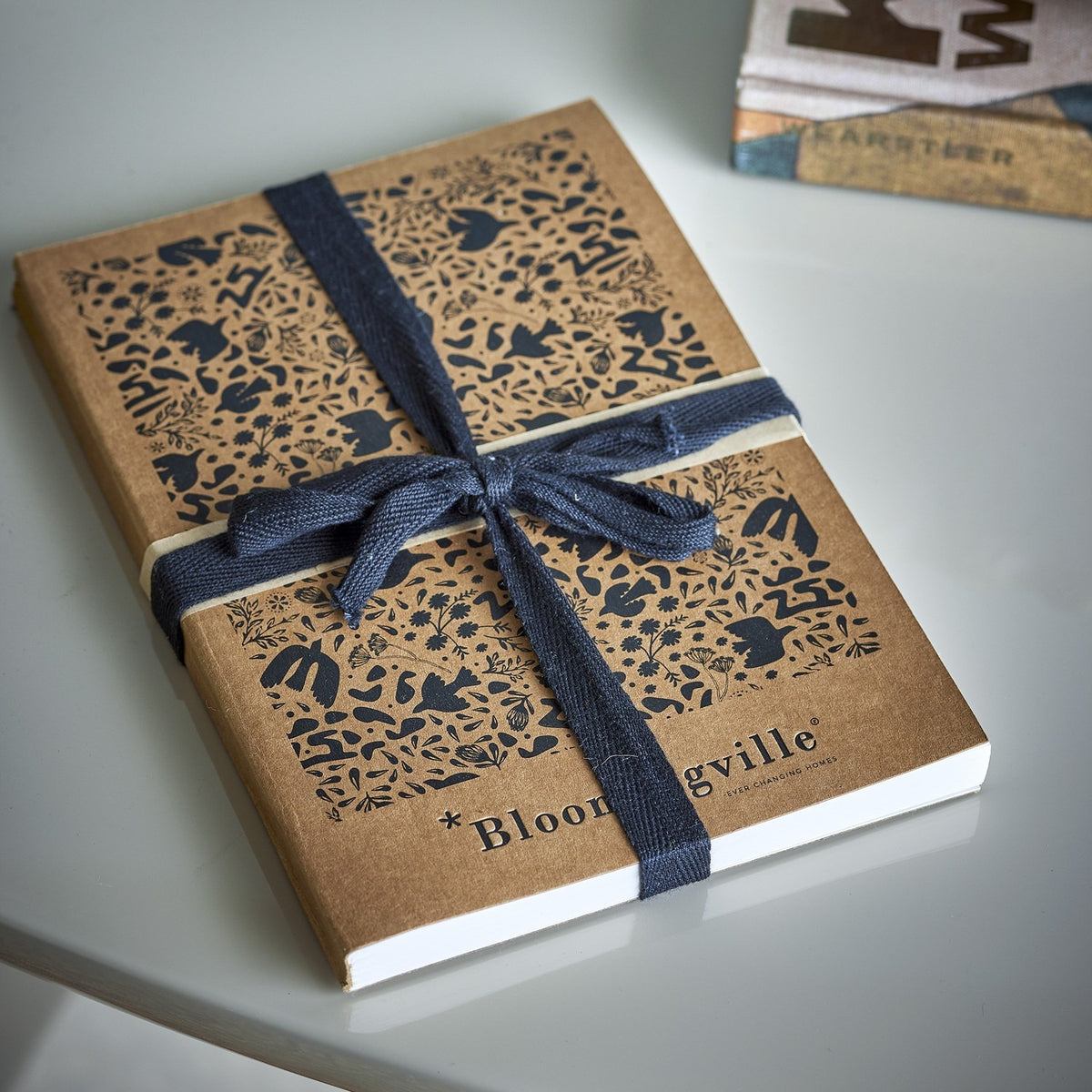 Anniversary Notebook in brown with a dense printed pattern of birds, flowers and leaves, tied with navy fabric ribbon, shown on a light surface.