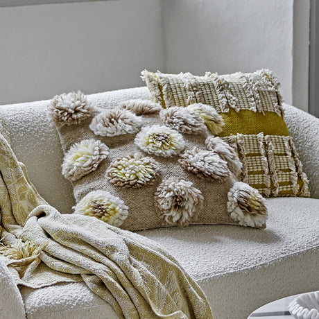 Somma Wool Cushion in Natural with tufted shaggy yarn rosettes on a woven base, shown on a boucle sofa beside patterned cushions and a throw.