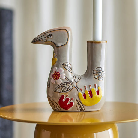 Vivielle Bird Stoneware Candle Holder in multicoloured floral design, bird-shaped with pointed beak and single candle tube, shown on a mustard side table with a white taper candle.