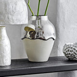 Silvio Stoneware Vase 26cm in Silver with glossy wavy-edged top and textured white lower body, shown on a dark shelf with green stems and decor nearby.