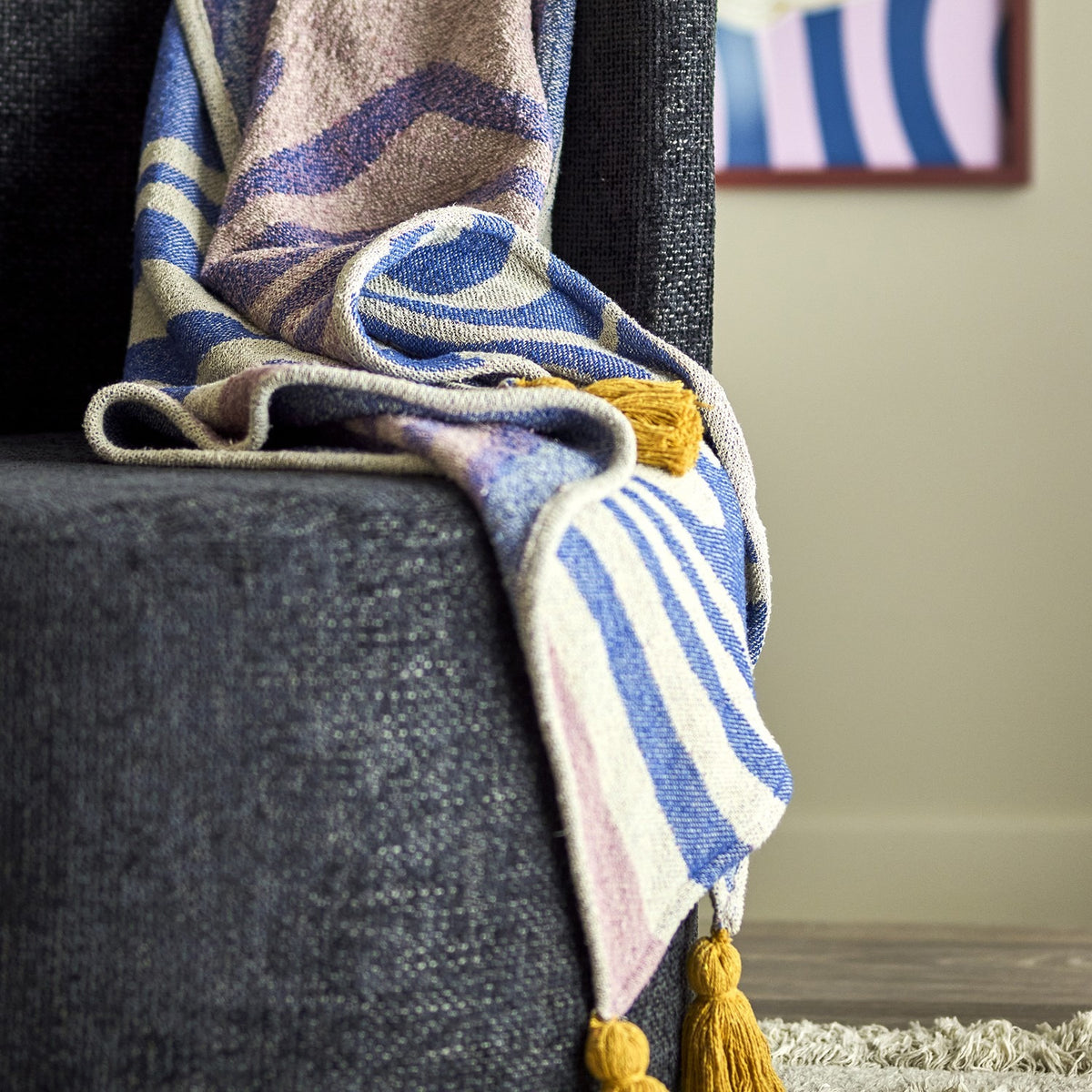 Arlie Throw in blue draped over a dark upholstered chair, showing a swirling pattern with striped sections and a mustard tassel at the corner.