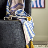 Arlie Throw in blue draped over a dark upholstered chair, showing a swirling pattern with striped sections and a mustard tassel at the corner.