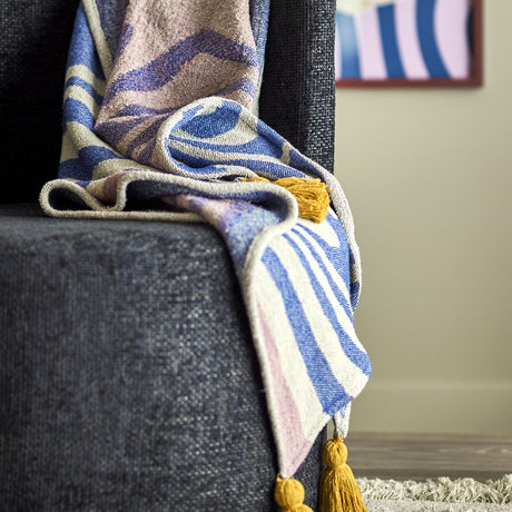 Arlie Throw in blue draped over a dark upholstered chair, showing a swirling pattern with striped sections and a mustard tassel at the corner.
