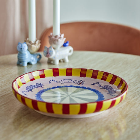 Missy Striped Stoneware Tray in multi, a round deep-sided ceramic dish with handpainted red and yellow rim stripes and a blue cat motif, shown on a table beside candle holders.