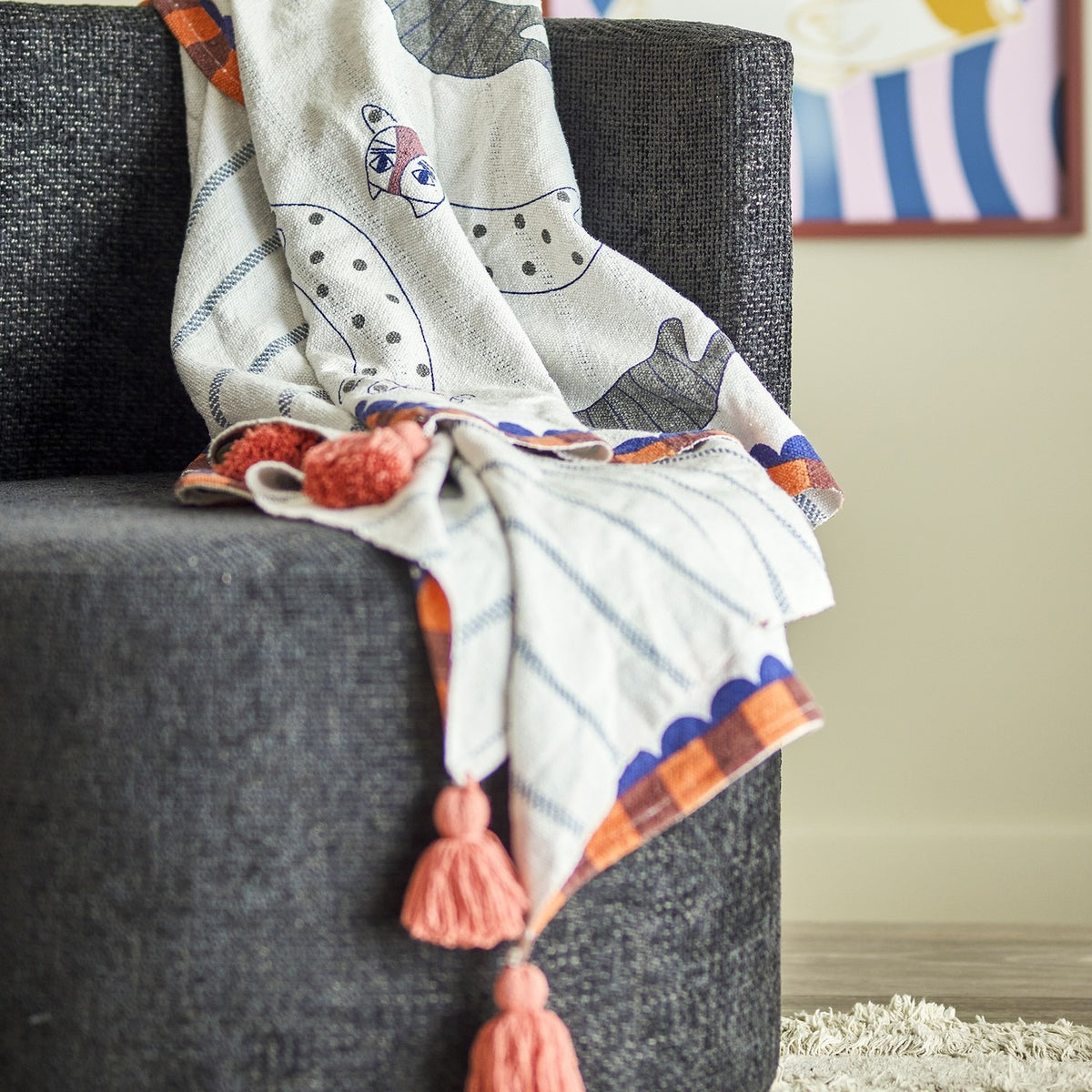 Multicoloured cotton-blend Missy Striped Throw draped over a dark grey armchair, featuring a whimsical cat print, spotted and striped bands, scalloped plaid border, and coral corner tassels.