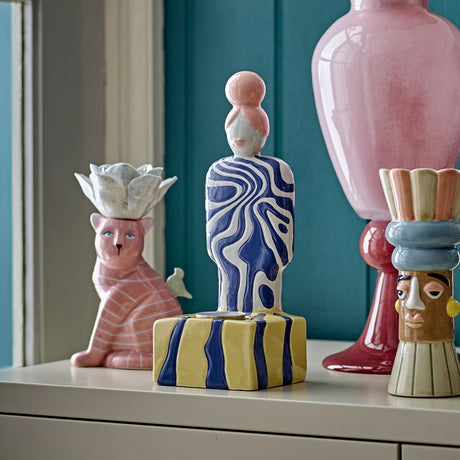 Priya Stoneware Tealight Holder in multicoloured ceramic, a figure with a bun and swirling navy-and-white pattern on a yellow striped base, shown among decorative vases on a shelf.