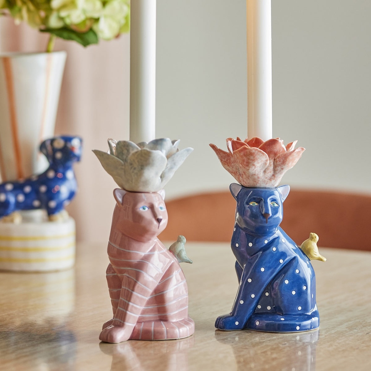 Eri Cat Stoneware Candle Holders, Set of 2, striped pink and dotted blue seated cats with floral candle cups and perched birds, shown on a table with candles lit.