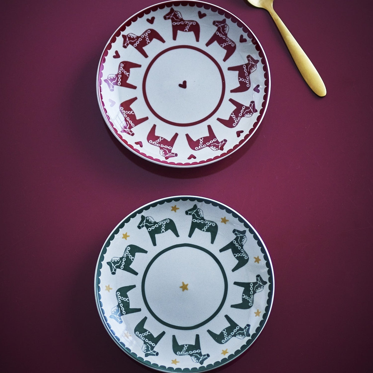 Siffer stoneware Christmas plates, set of 2, scalloped rims, red plate with horses and hearts, green plate with horses and stars, shown with a gold spoon.
