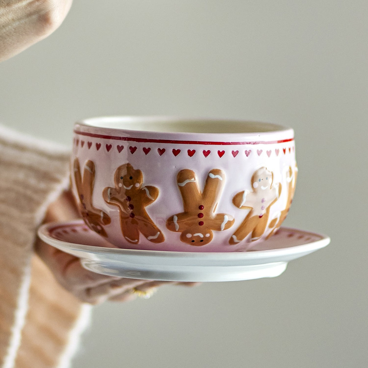 Hand holding a pink stoneware bowl from the Binna Heart Christmas set, featuring embossed gingerbread figures and a heart border, shown with a matching saucer.