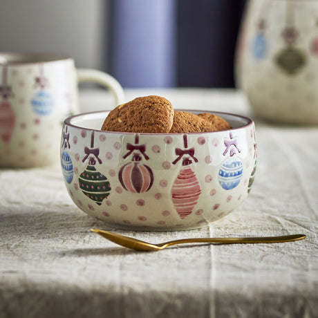 Ornamentia stoneware bowl with embossed Christmas bauble, bow and polka dot design in pink, blue and green, filled with biscuits beside a matching mug and gold spoon.