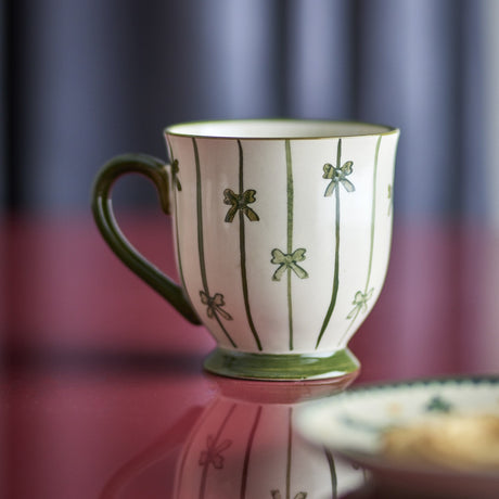 Hope Striped Stoneware Christmas Mug with cream glaze, green vertical stripes, hand-painted bows, curved handle and footed base, set against a dark striped background.