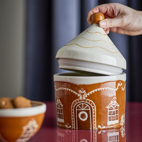Gina Stoneware Christmas Storage Jar 1150ml in brown, shaped like a gingerbread house with piped icing-style windows, door wreath and a pointed roof lid with round knob, shown lifted open by hand.