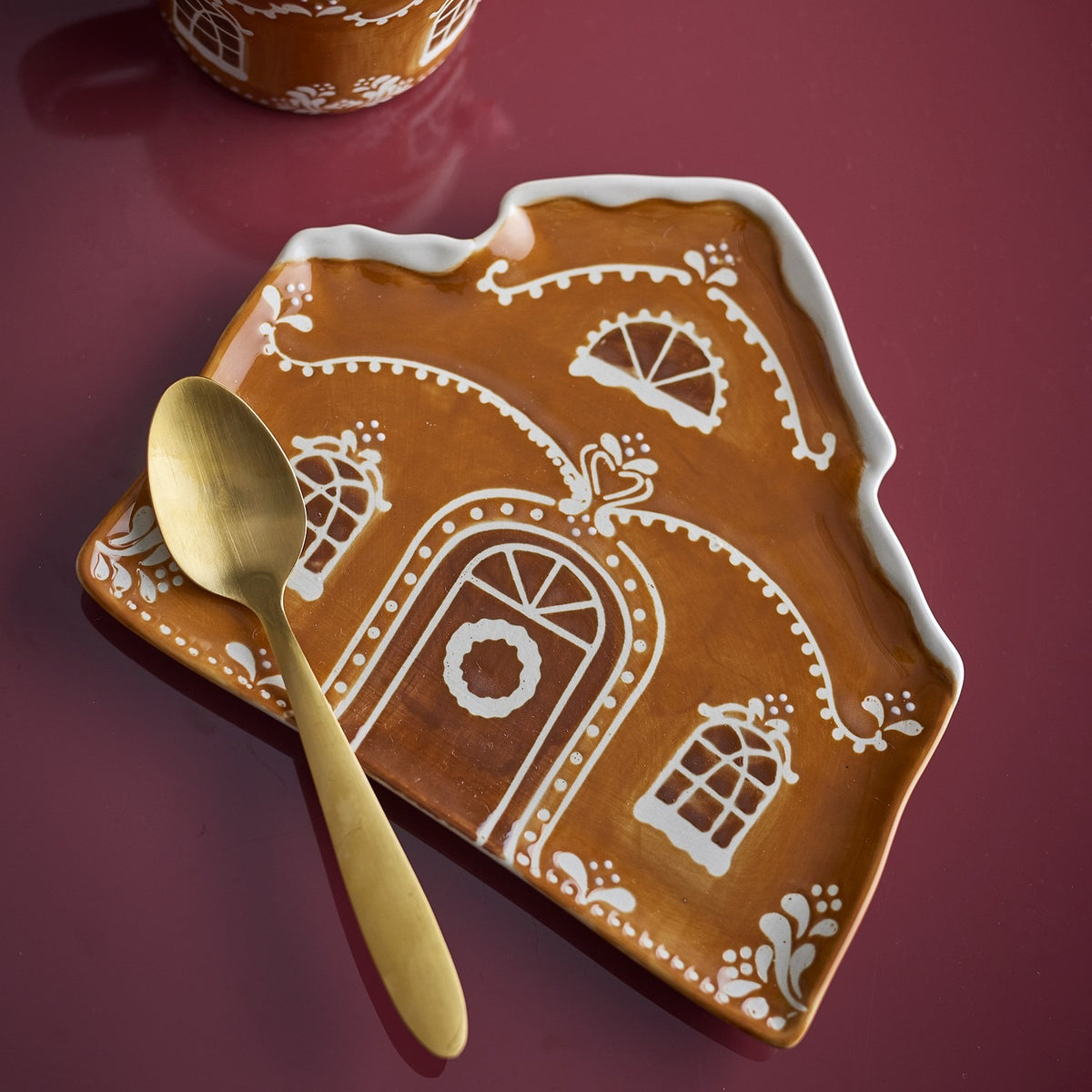 Gina House stoneware Christmas platter in brown, shaped like a gingerbread house with hand-painted door, wreath and windows, shown with a gold spoon on a dark red surface.