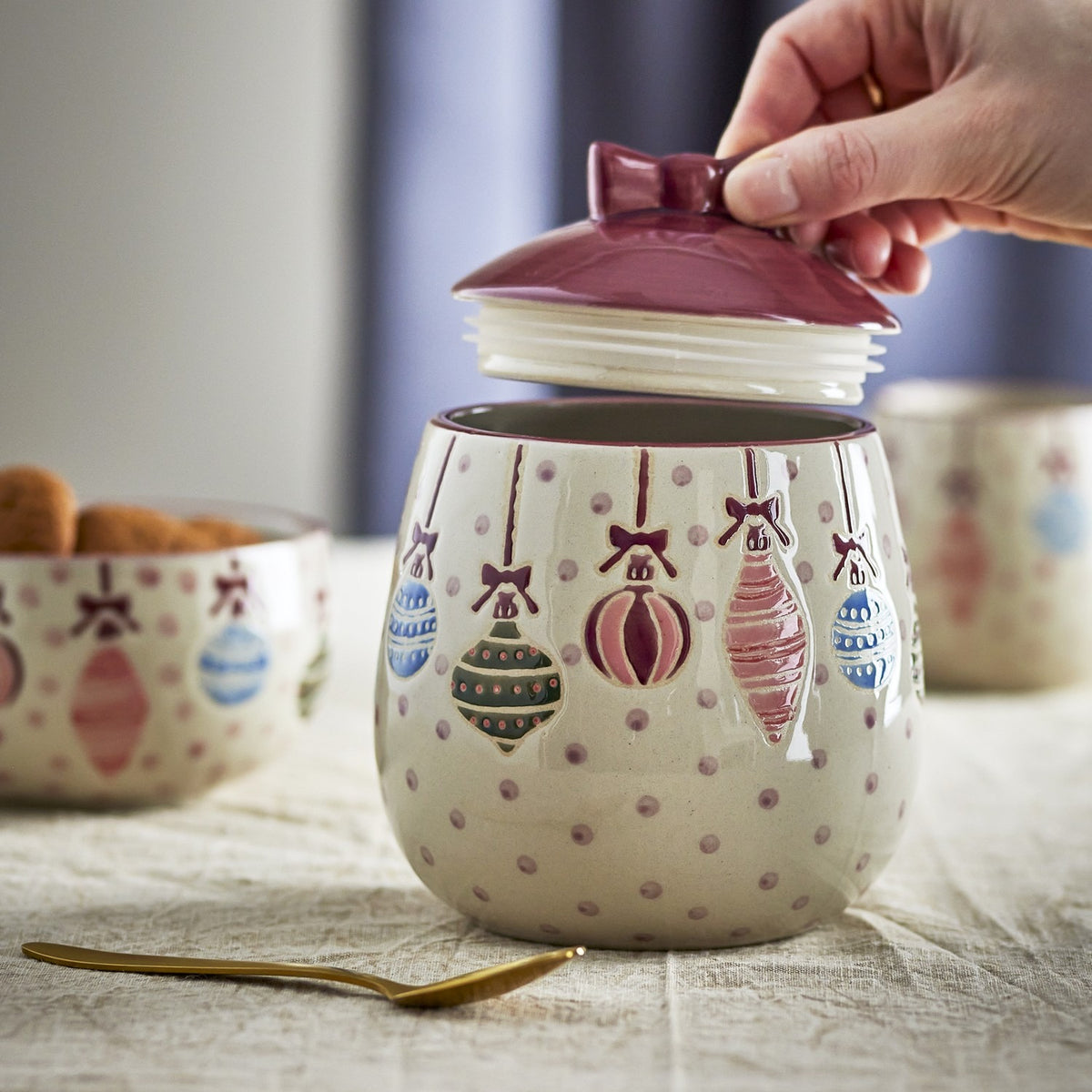 Ornamentia Dotted Christmas Storage Jar in cream stoneware with hand-painted hanging baubles, being opened by hand to reveal a plum-glazed lid with a bow-shaped knob.