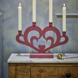 Salia Heart Advent Candle Holder in red MDF, a heart-shaped cut-out design with scrolls and four lit candles, displayed on a marble-top wooden cabinet.