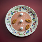 Caramella Dotted Stoneware Christmas Dinner Plate with multicoloured wrapped-sweet rim pattern and dotted grid centre, shown with cookies and sugared balls on a dark red background.