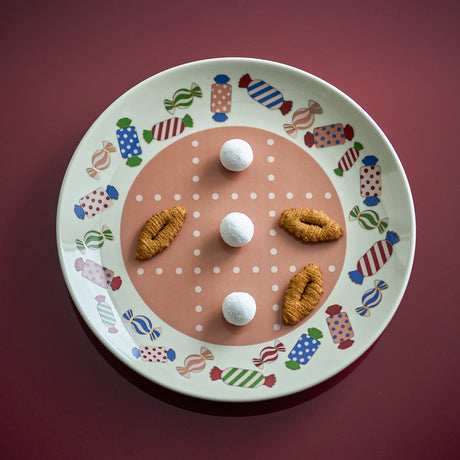 Caramella Dotted Stoneware Christmas Dinner Plate with multicoloured wrapped-sweet rim pattern and dotted grid centre, shown with cookies and sugared balls on a dark red background.