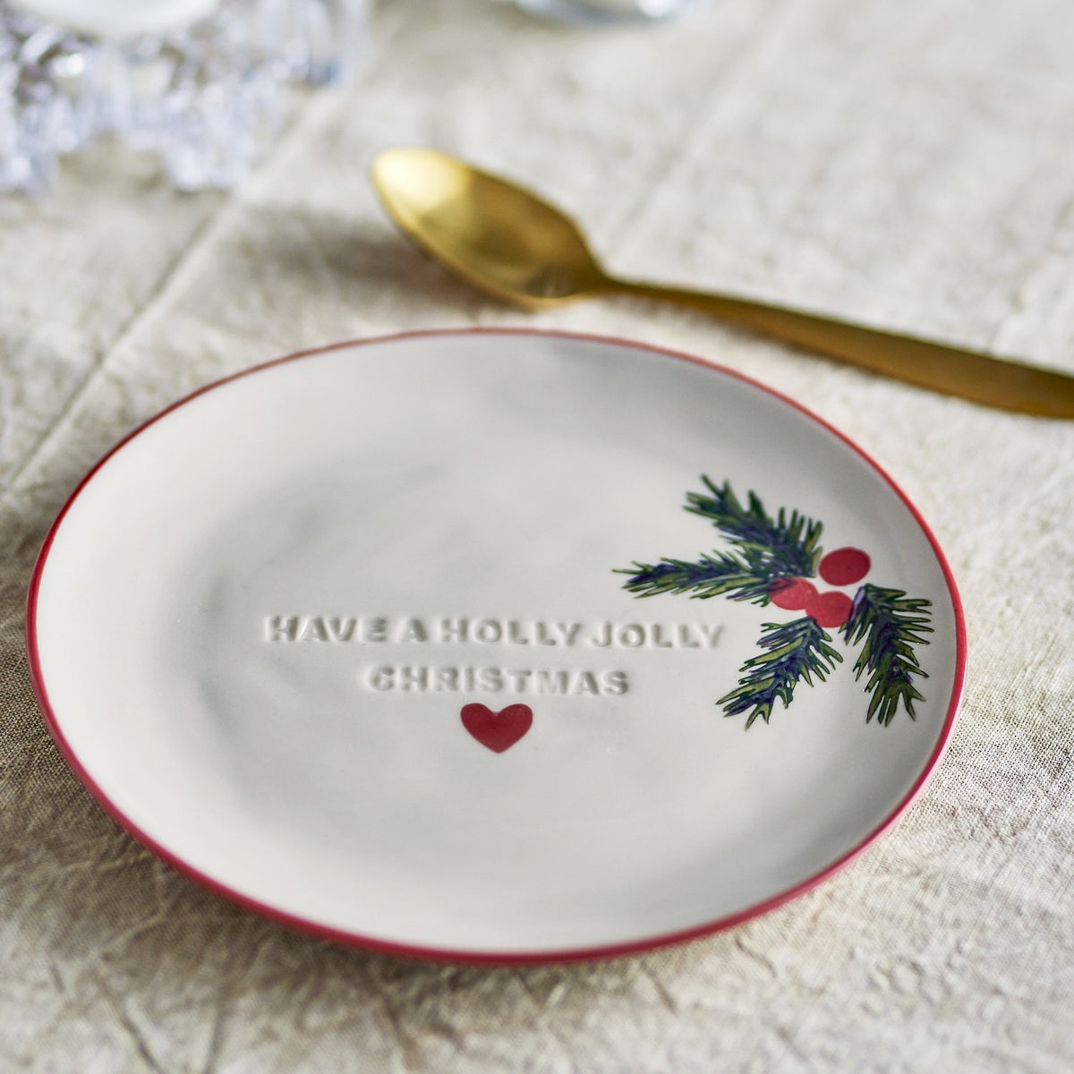 Cream stoneware Christmas side plate with red rim, embossed "Have a Holly Jolly Christmas" text, a red heart, and a painted holly sprig, styled with a gold spoon on linen.