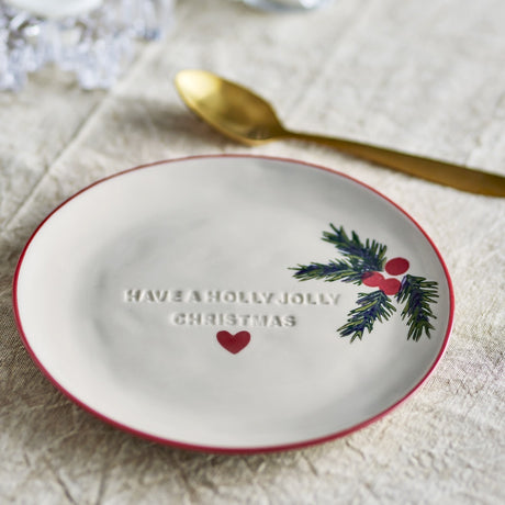 Cream stoneware Christmas side plate with red rim, embossed "Have a Holly Jolly Christmas" text, a red heart, and a painted holly sprig, styled with a gold spoon on linen.