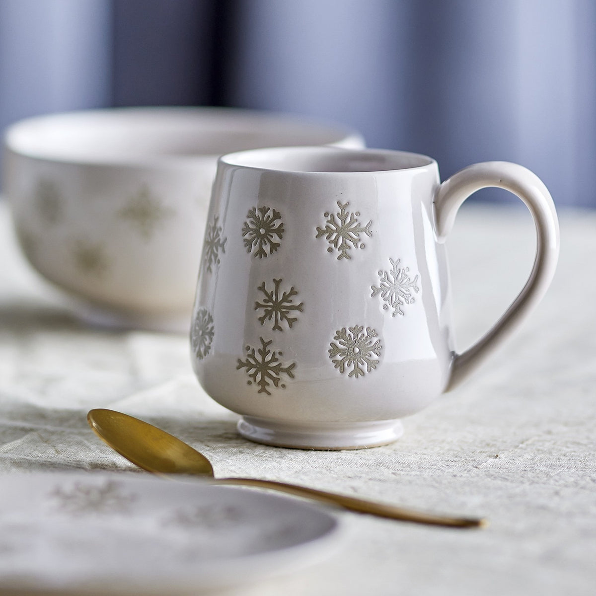 Cream stoneware Christmas mug with hand-painted snowflake pattern and plain curved handle, shown with a matching bowl, saucer, and gold spoon.