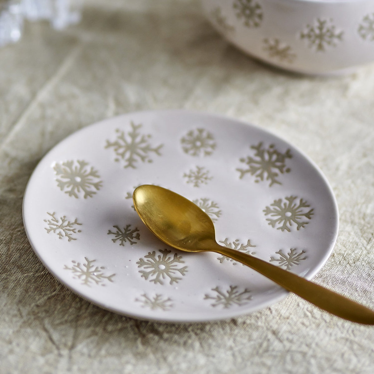 Beige stoneware side plate from the Snowie Snowflake Christmas set, scattered with varied snowflake designs, shown with a gold spoon on a textured neutral tablecloth.