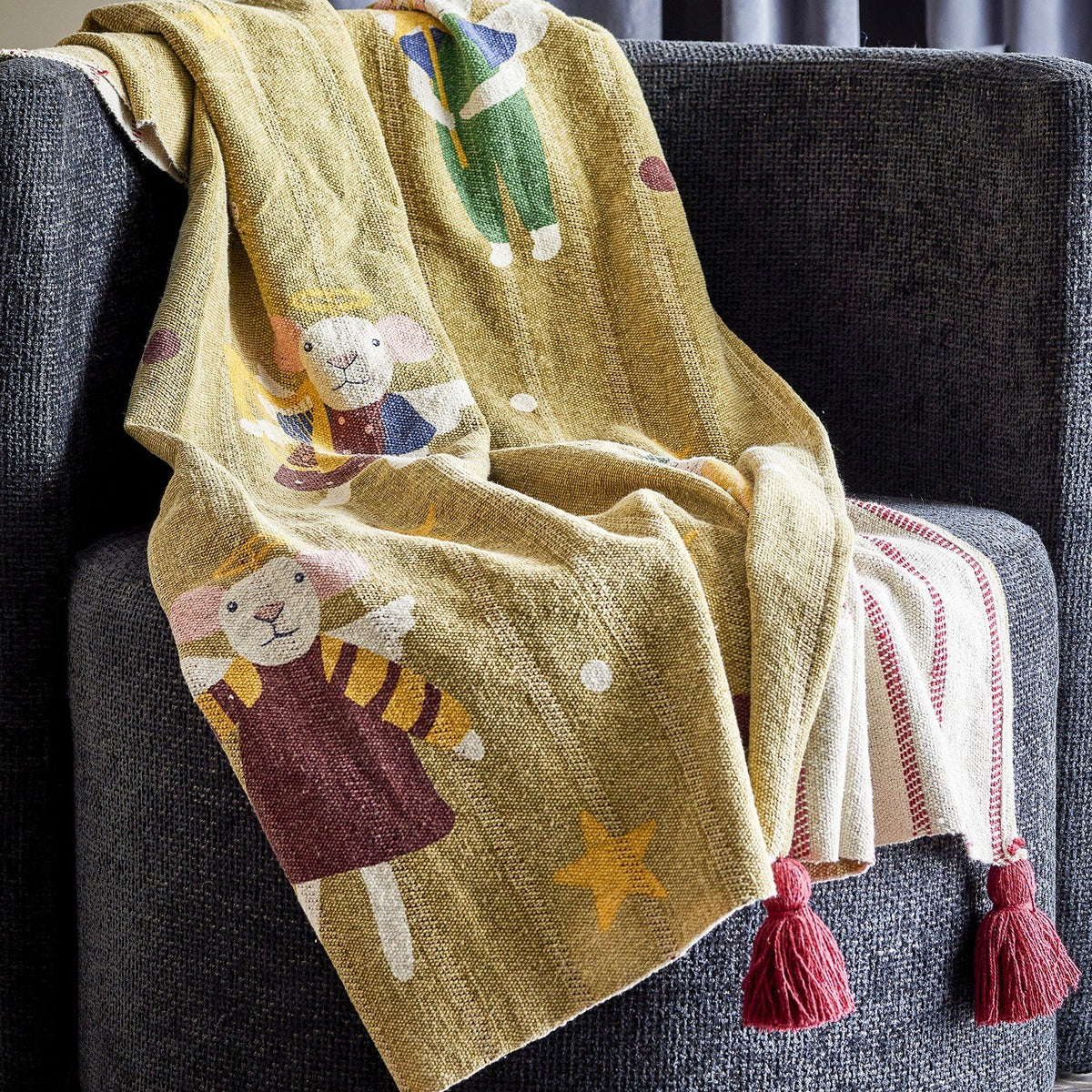 Peo Striped Christmas Throw in green, draped over a grey armchair, showing mice-in-angel-costume motifs with stars on one side and a red-striped reverse with tasselled corners.