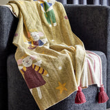 Peo Striped Christmas Throw in green, draped over a grey armchair, showing mice-in-angel-costume motifs with stars on one side and a red-striped reverse with tasselled corners.