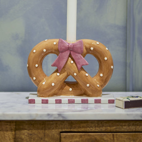 Pretzie pretzel-shaped ceramic candle holder in brown with white dots and a pink bow, on a striped base with a white taper candle, shown on a marble surface.