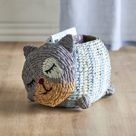 Mille Water Hyacinth Basket shaped like a sleeping cat with woven grey, cream and tan body, embroidered eyes, stubby feet and curled tail, shown on a wood floor holding magazines.