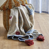 Mille Cotton Throw in beige draped over a woven basket, showing a printed cat and yarn-ball scene, blue striped reverse, and burgundy tasselled corners.