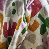 Filur Cotton Throw in Multi, draped fabric with abstract green, mustard, pink and burgundy shapes on a cream woven background.