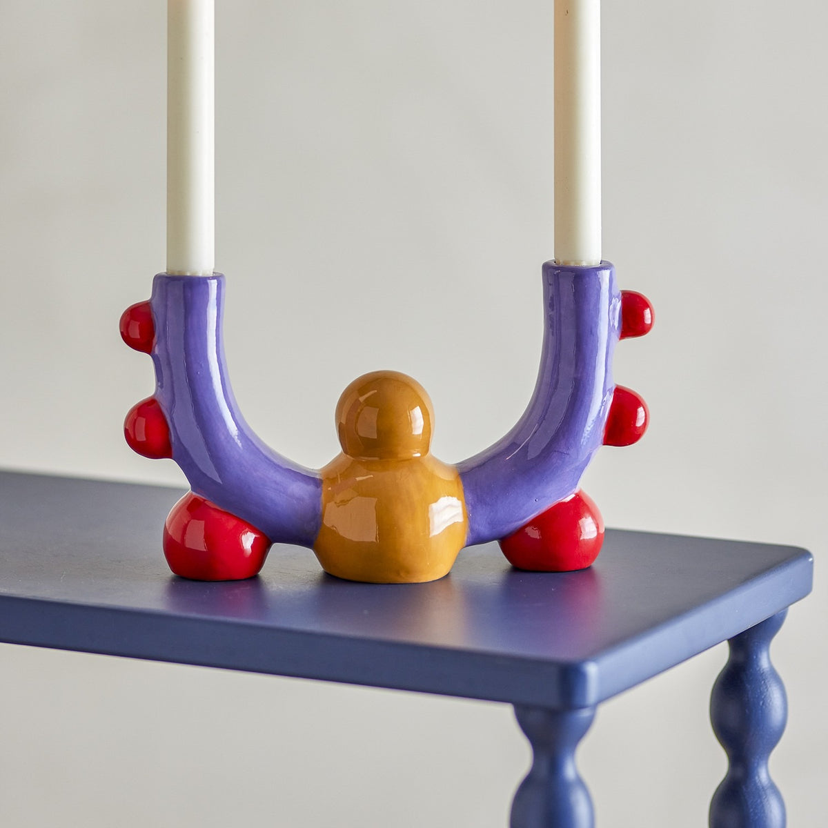 Edivia Stoneware Candle Holder in multicoloured ceramic, with purple U-shaped arms, red ball details, and an amber rounded base, holding two white dinner candles on a dark blue table.