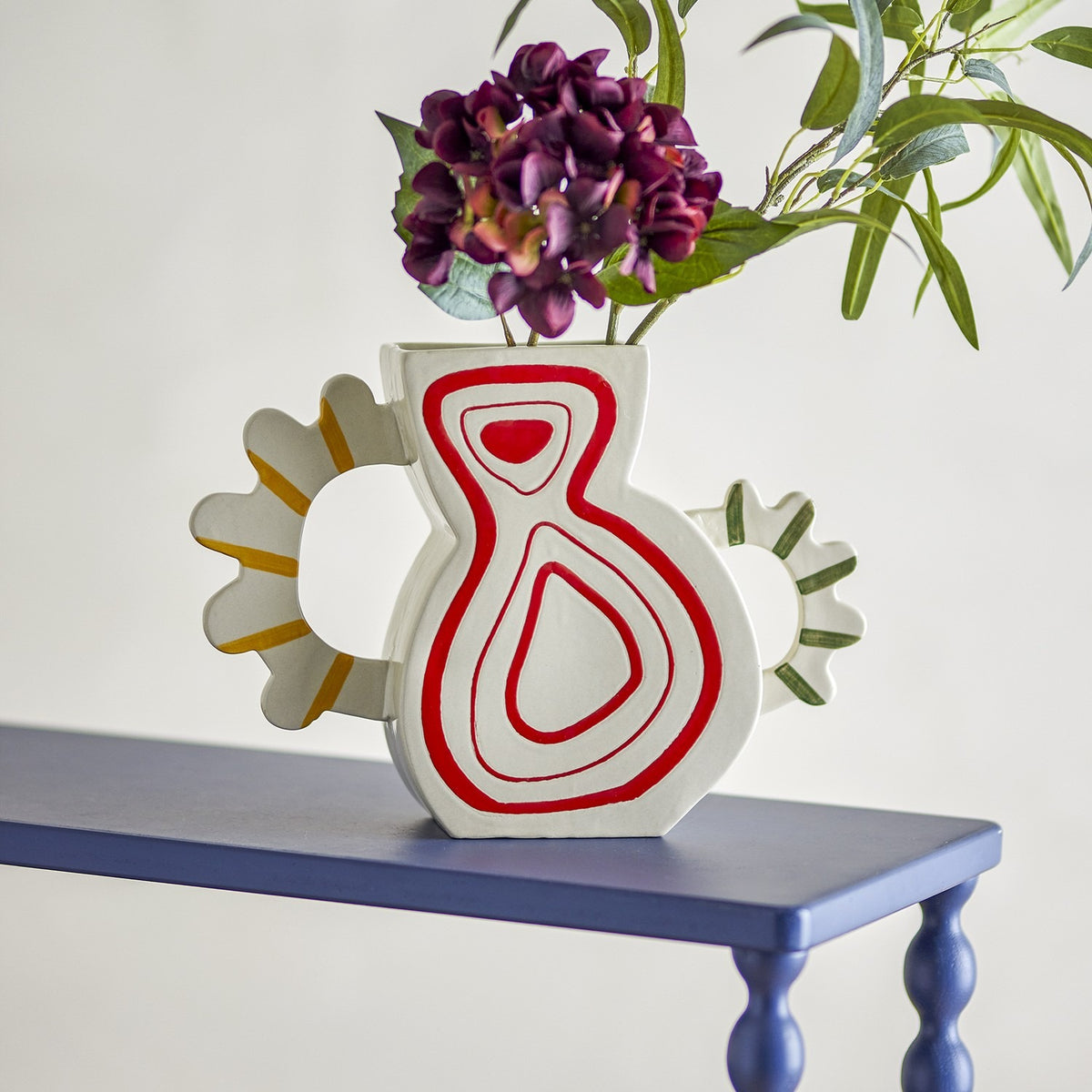 Samila Striped Stoneware Vase 26cm in white with red concentric droplet motif, flat flask-shaped body and scalloped ring handles, holding purple flowers on a blue table.