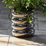 Shine stoneware plant pot with saucer in silver, stacked rounded rings creating a ribbed, reflective tiered design, shown planted on a wooden floor.